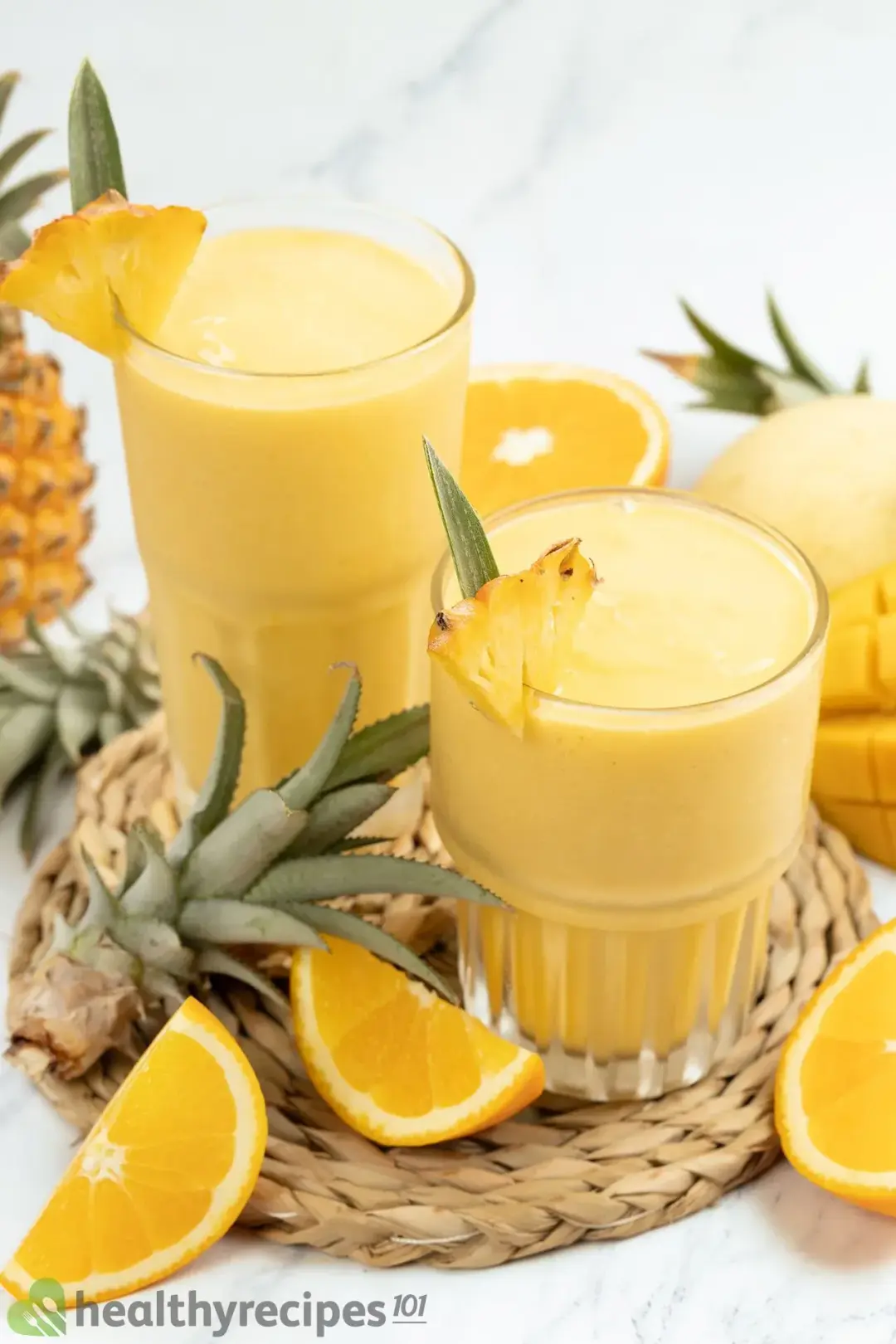 Two glasses of orange smoothie with orange wedges on the side and pineapple tops as garnishes