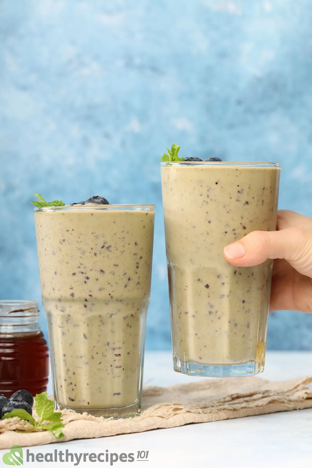 A hand picking up a glass of blueberry avocado smoothie laid on a wooden board near an identical glass, mint leaves, and a jar of honey.