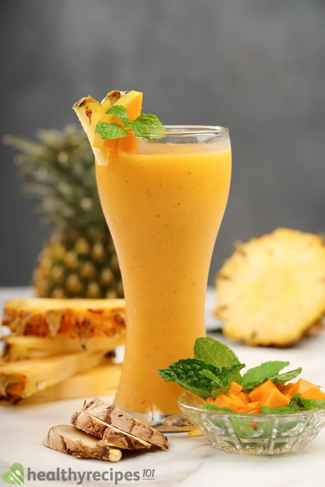 A tall glass of Mango Ginger Smoothie placed near ginger, mint leaves, and a small bowl of mangoes with pinapples in the back.