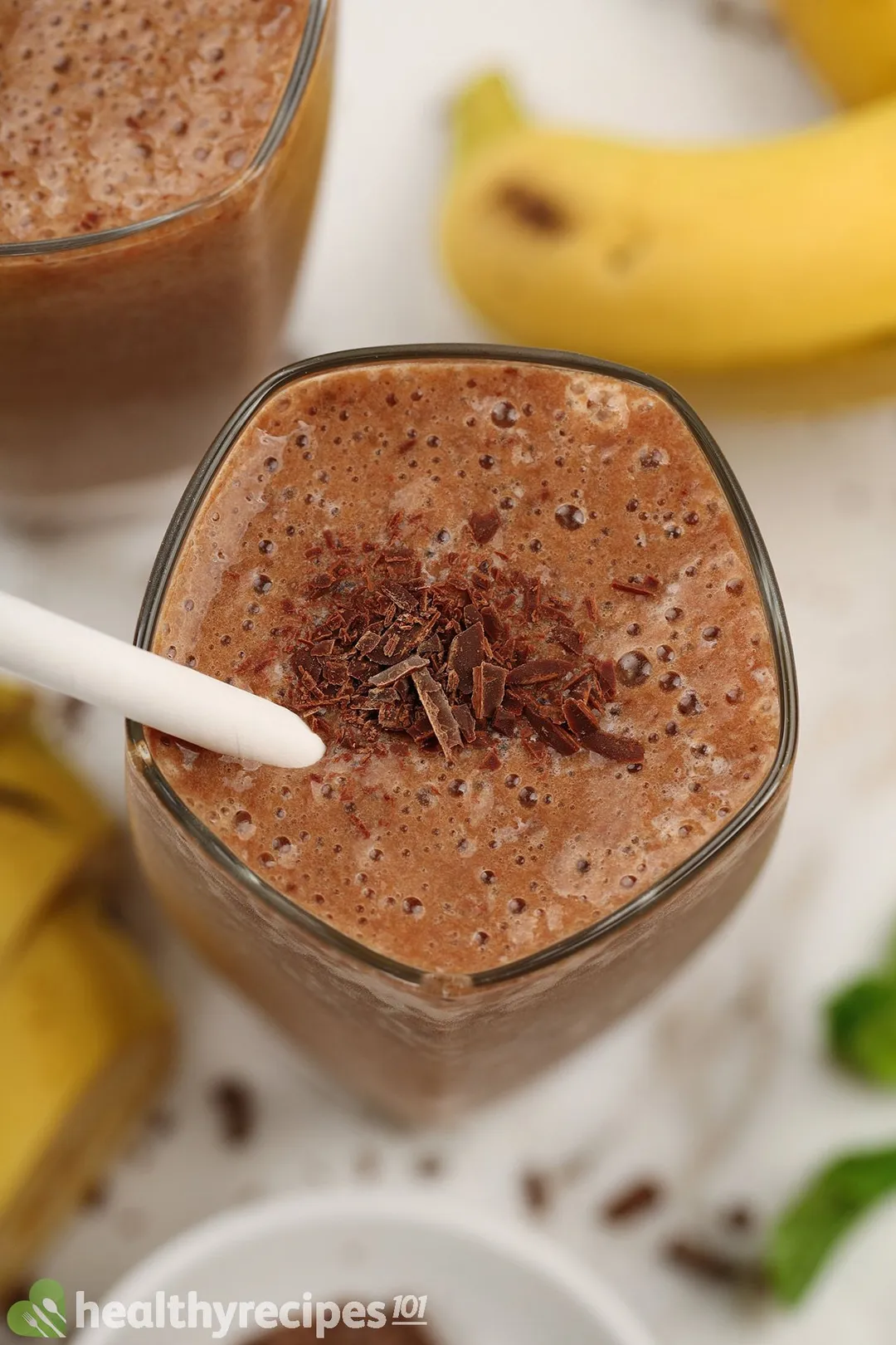 A high-angle shot of a glass of chocolate banana smoothie with yellow bananas in the blurred bottom background.