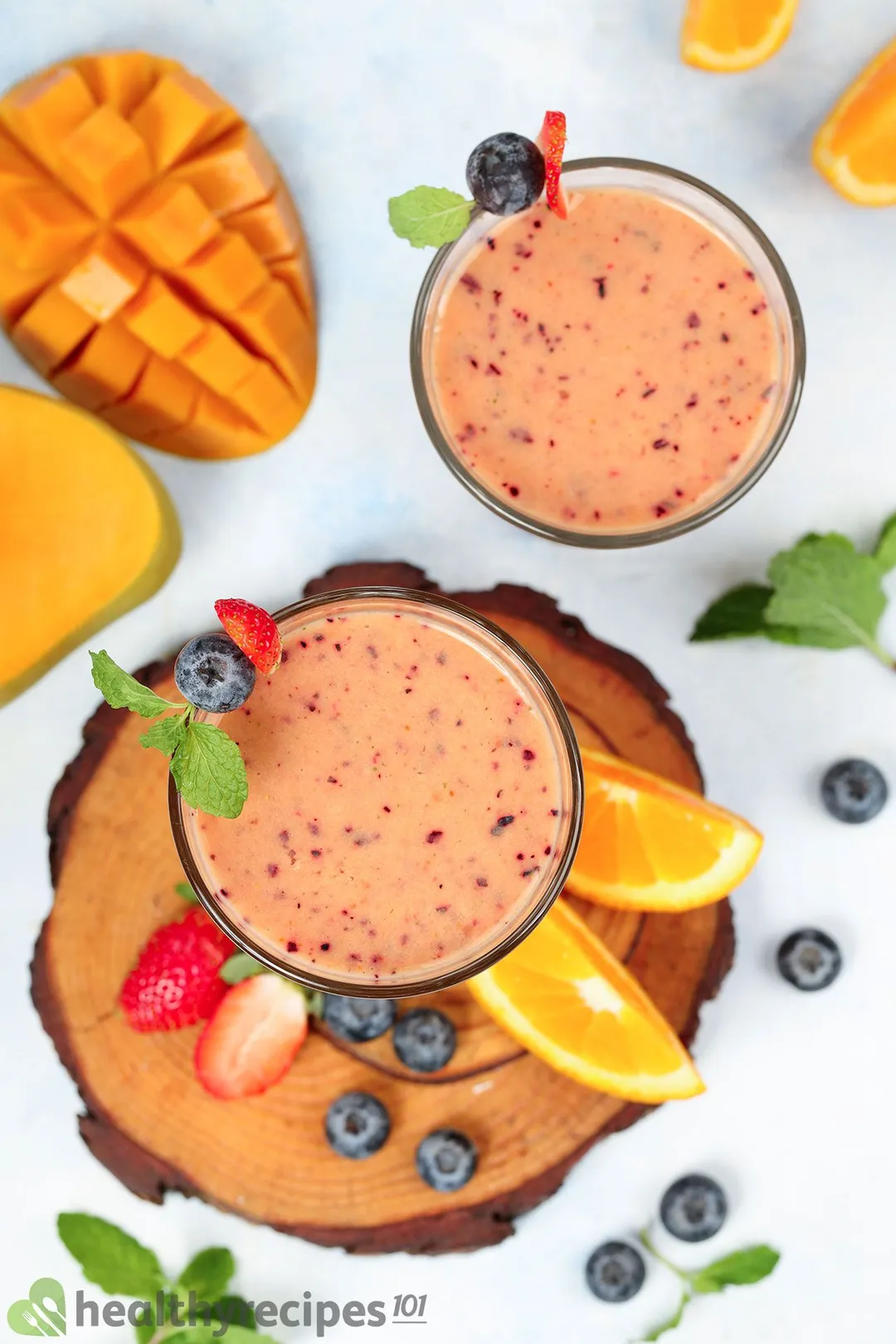 A high-angle shot of two glasses of Mango Berry Smoothie placed on a wooden board and surrounded by blueberries, orange wedges, scored mangoes, and mint leaves.