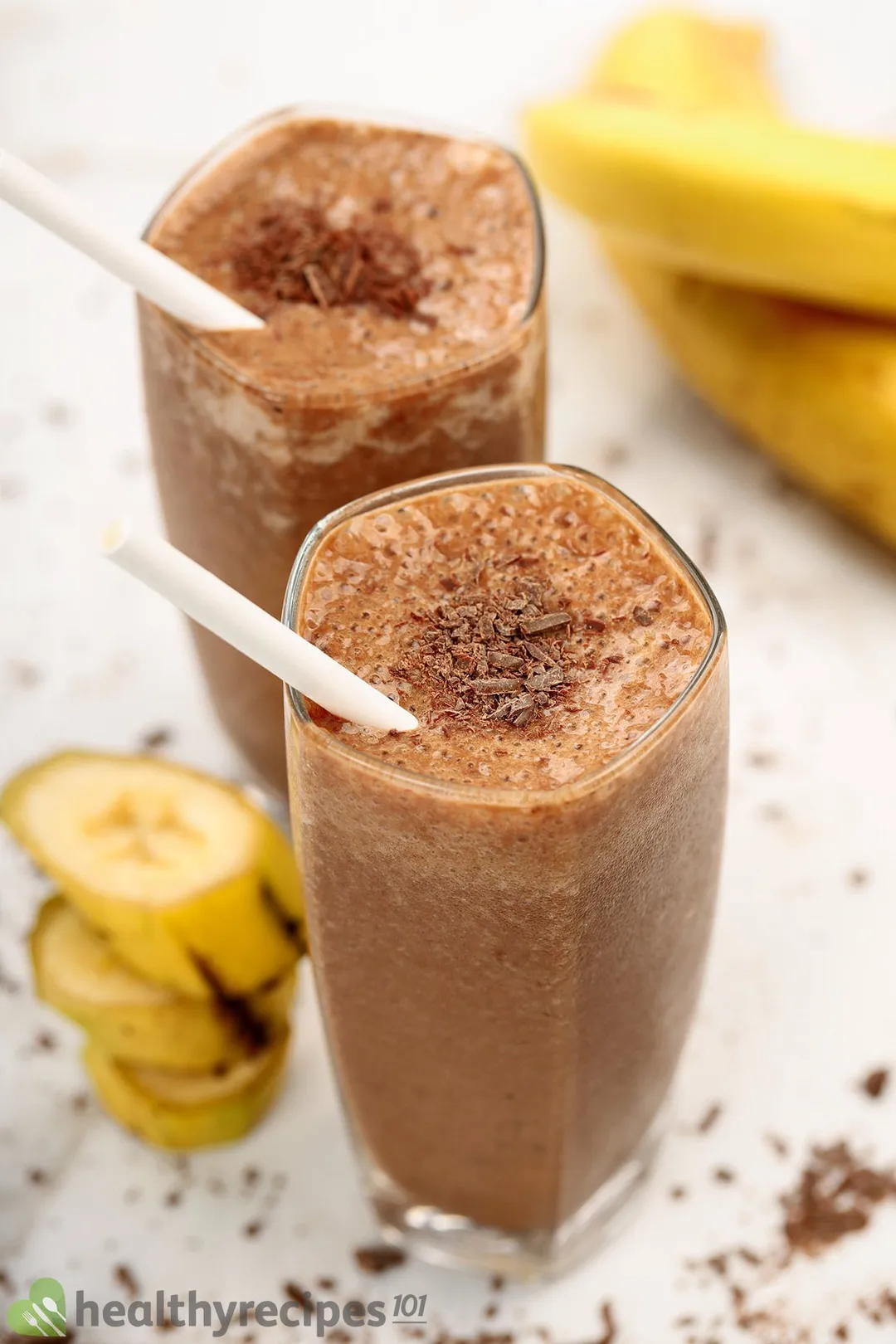 Two glasses of chocolate banana smoothies placed near unpeeled yellow bananas.