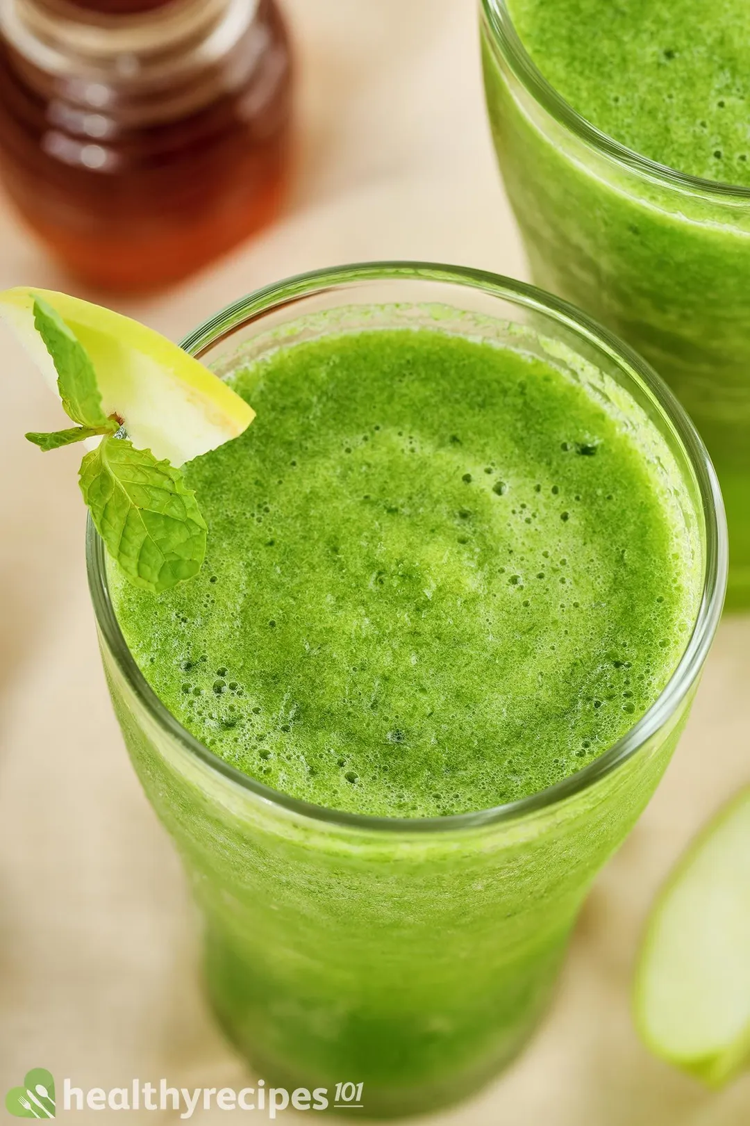 A high-angle shot of a vibrant green glass of sour apple smoothie. Next to it is a similar glass and a small jar of honey.