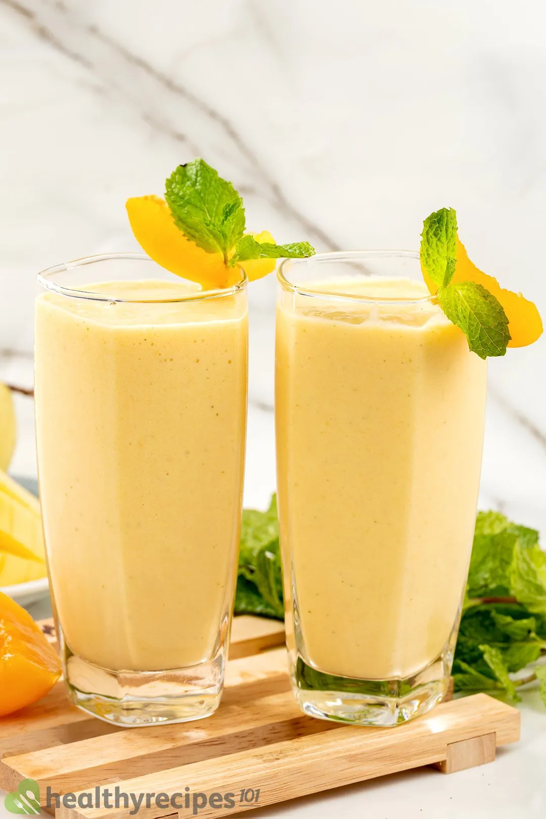 Two glasses of peach smoothie placed on a wooden board with some mint leaves and peach slices in the back.