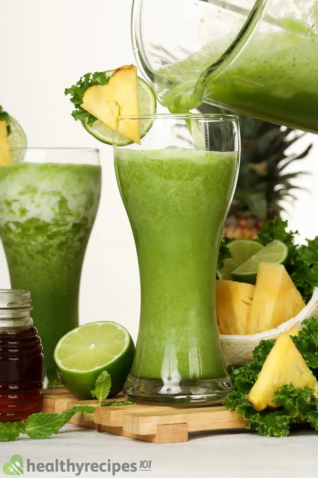 A pitcher of smoothie being poured into a glass decorated with a slice of pineapple alongside a glass of green mojito smoothie and ingredients of the smoothie.