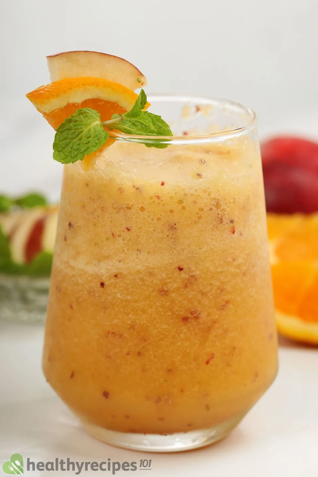 A glass of apple orange smoothie garnished with a small slice of orange, a mint julep, and a small slice of apple.