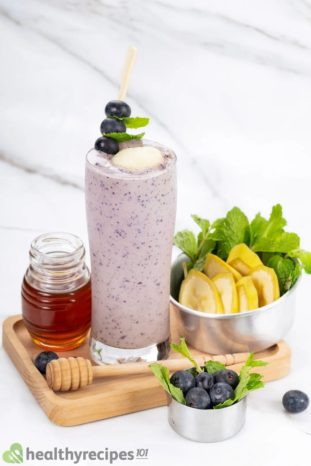 A tall glass of blueberry banana smoothie placed on a wooden board and surrounded by small bowls of bananas, blueberries, and mint leaves, and a small jar of honey.