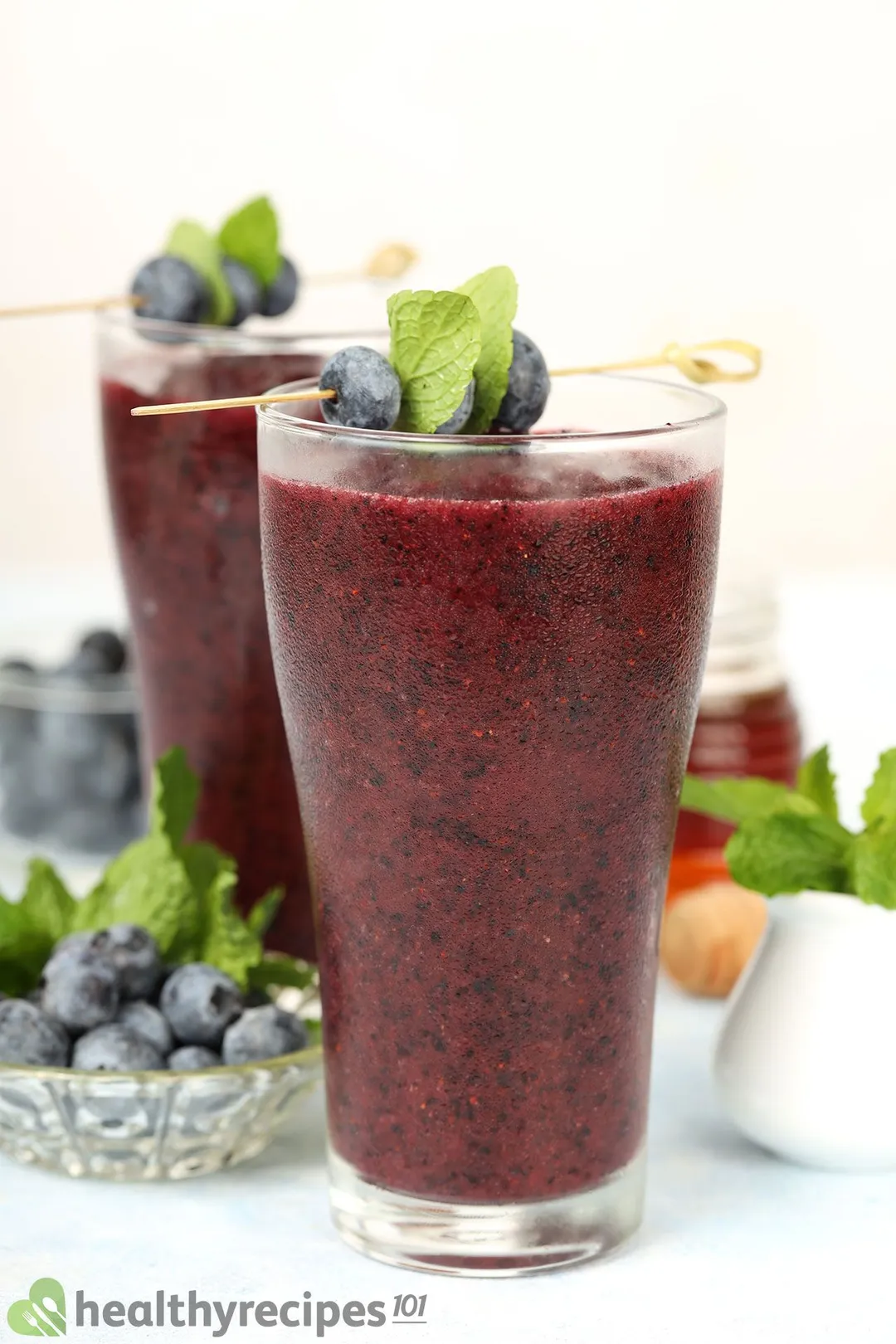 Two glasses of blueberry smoothie garnished with blueberry skewers and placed near small plates of blueberries and a small jar of honey.