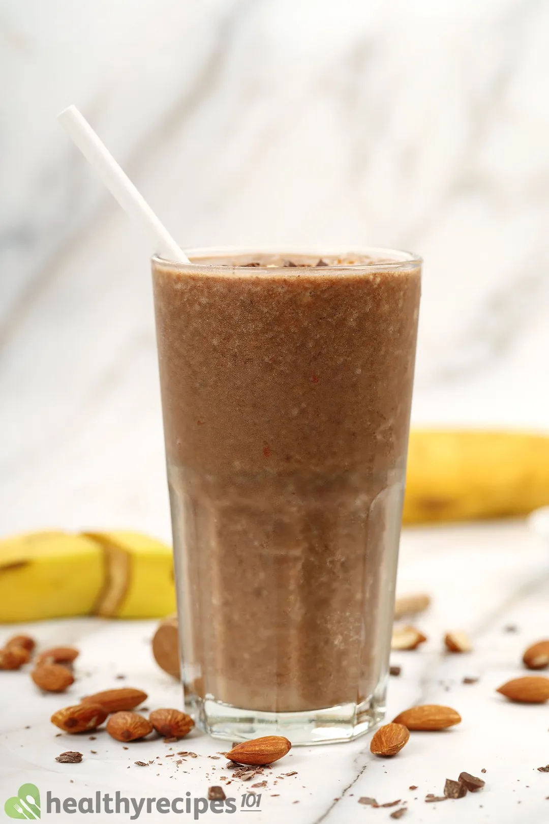 A glass of chocolate peanut butter banana placed on a marbled surface scattered with unpeeled bananas and almonds.