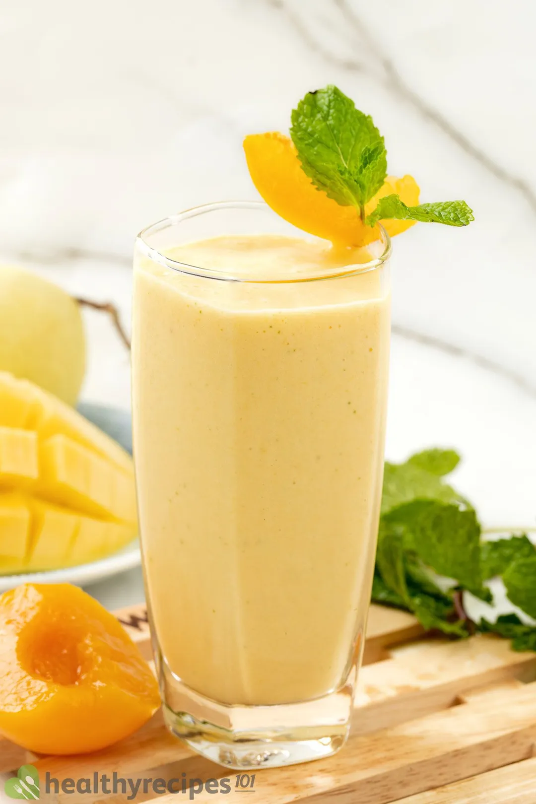 A glass of peach smoothie placed on a wooden board with a peach, sliced mango, and mint leaves nearby.