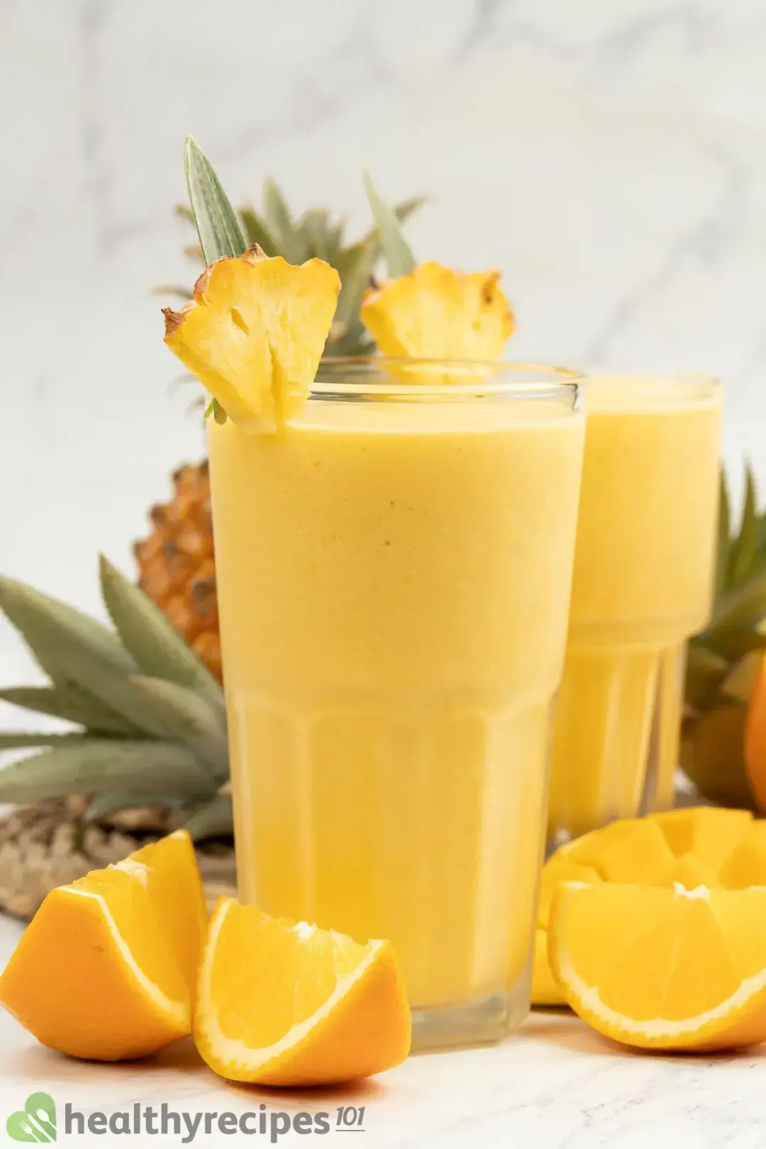Two tall glasses of orange smoothie with orange wedges on the side and pineapple wedges on the rim