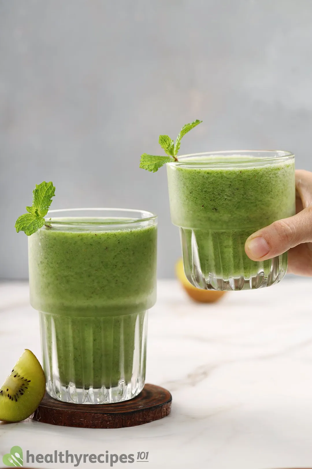 A hand holding up a small glass of apple kale smoothie next to a taller glass placed on a wooden coaster.