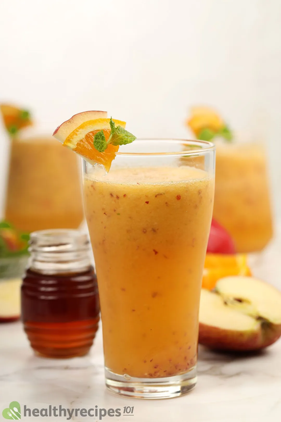 A glass of apple orange smoothie laid near a small jar of honey and half an apple with two other similar glasses in the blurred background.