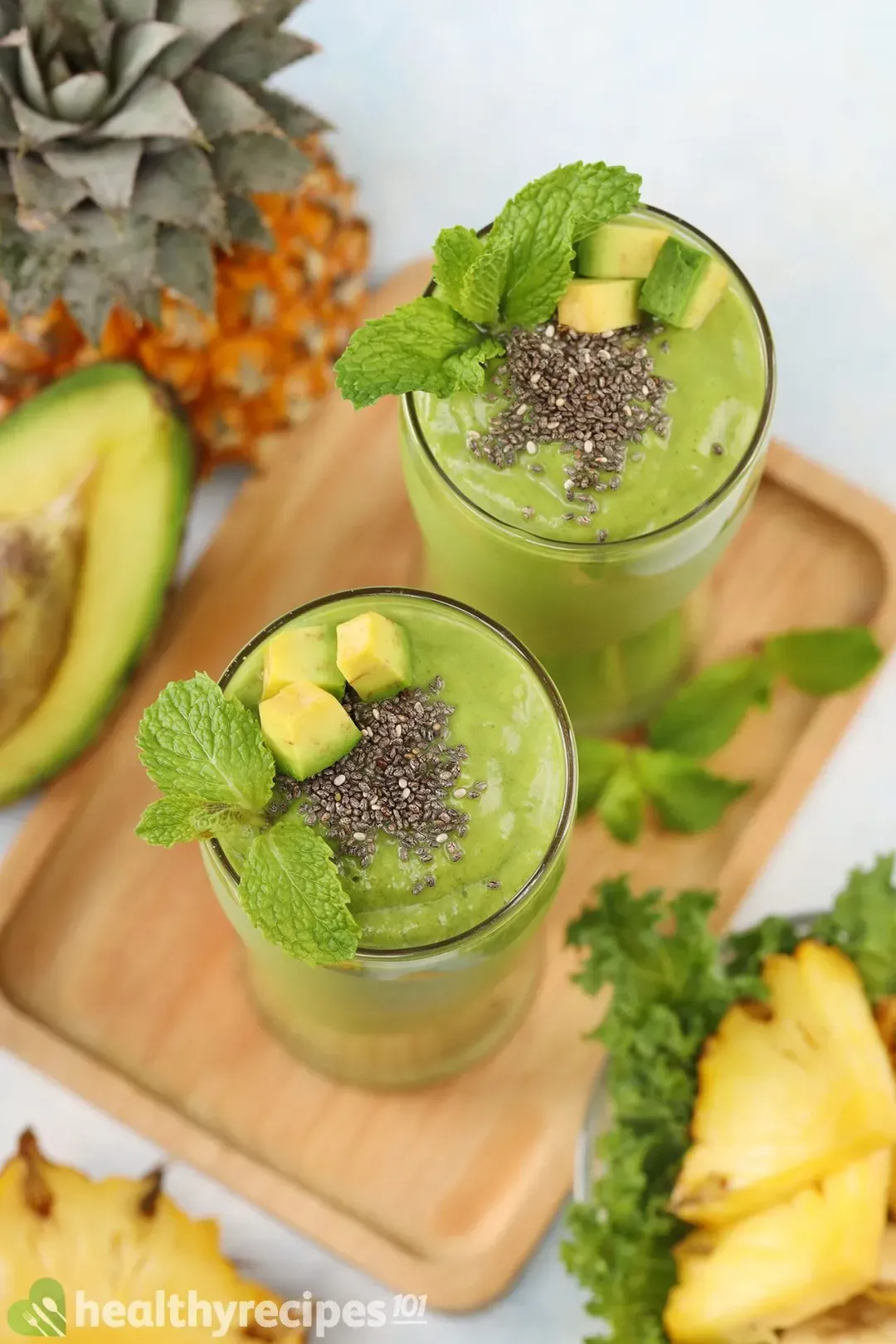two glasses of avocado green smoothie on a wooden tray with pineapple wedges and avocado slices and chia seed on top
