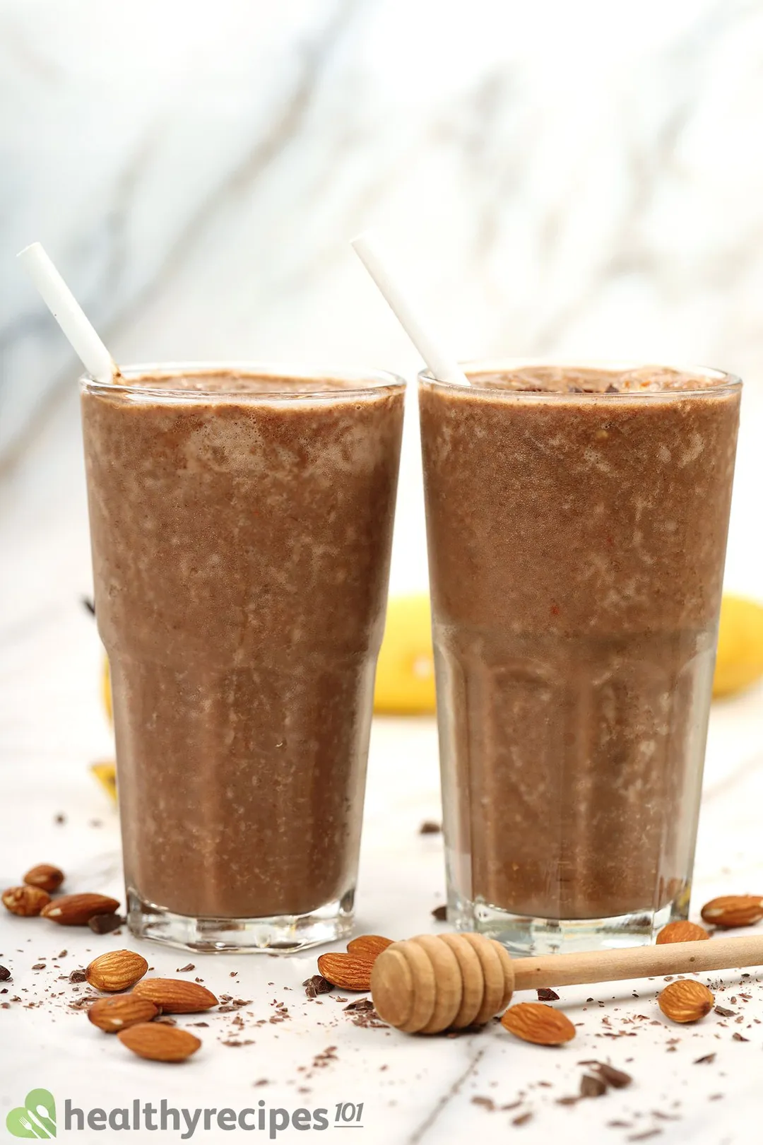Two glasses of chocolate peanut butter banana smoothie surrounded with almonds.