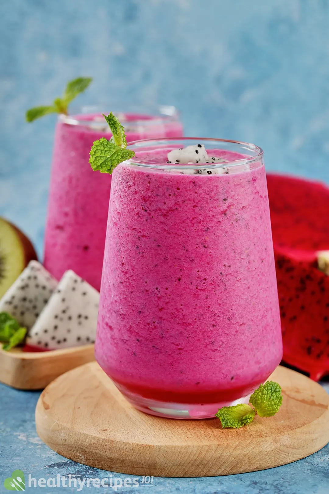 A glass of Dragon Fruit Smoothie placed on a round wooden board with a similar glass and a few dragon fruit slices in the back.