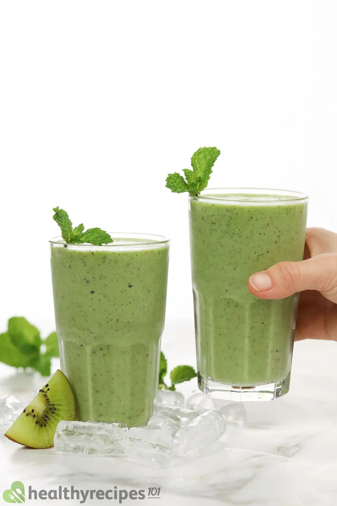 A hand picking up a glass of kiwi smoothie placed next to an identical glass. They're both placed on a white cloth with a slice of green kiwi and mint leaves near by.