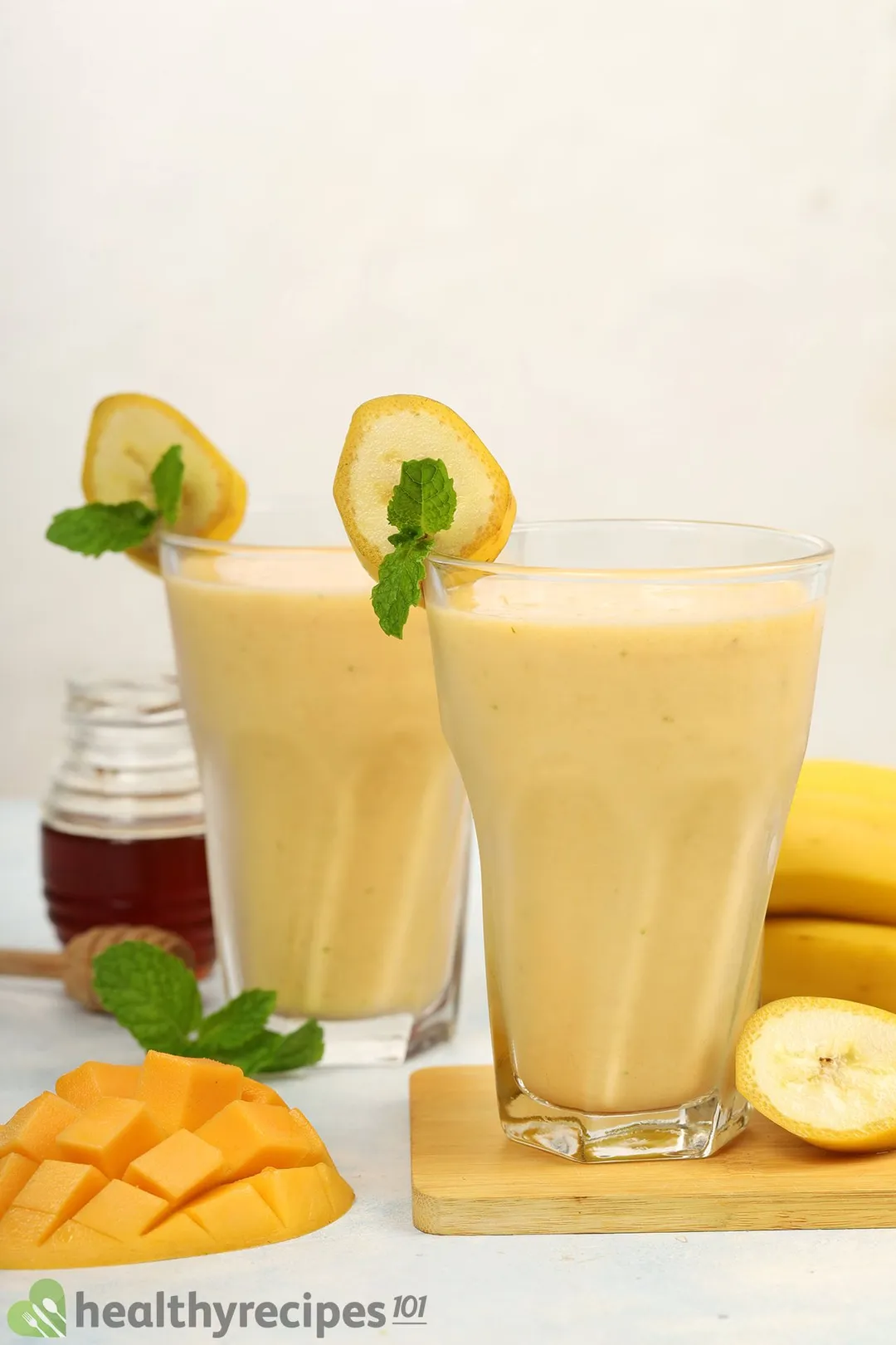 Two glasses of Mango Banana Smoothie placed near a scored mango, sliced bananas, mint leaves, and a small jar of honey.