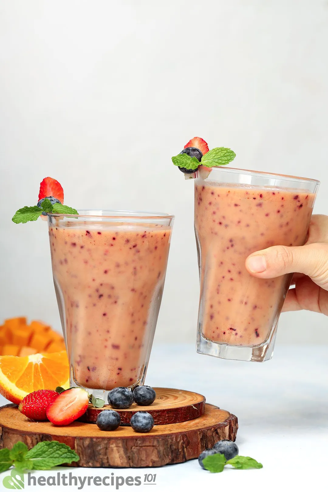 Two glasses of Mango Berry Smoothie placed on a wooden board near strawberries, blueberries, and half an orange. A hand is picking one of them up.