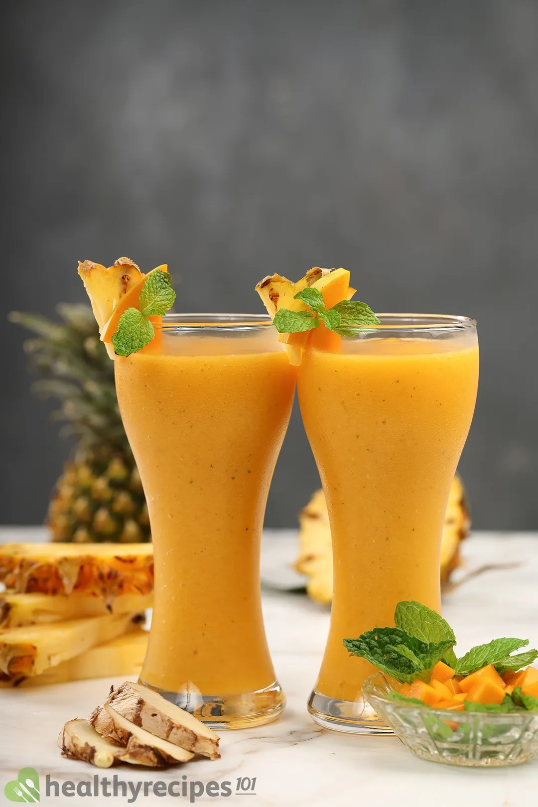 Two glasses of Mango Ginger Smoothie placed near ginger, mangoes, and pinapples.