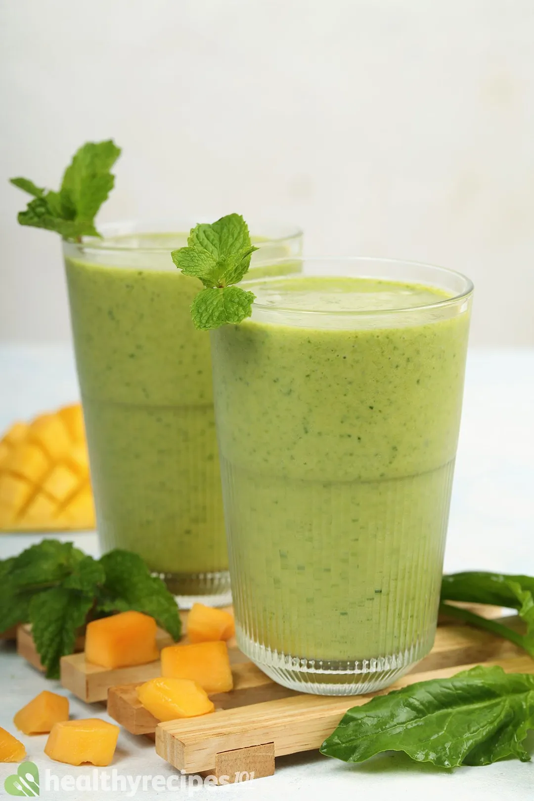 Two glasses of Mango Spinach Smoothie placed on a wooden board near fresh spinach and cubed mangoes.