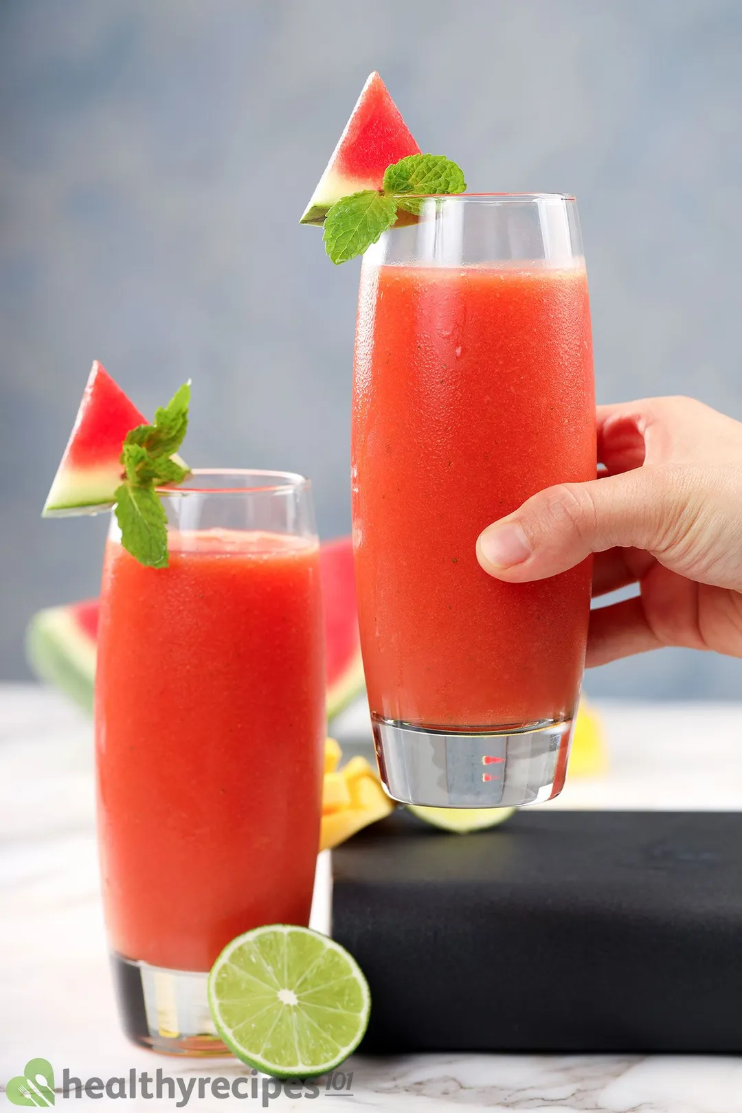 A hand picking up a glass of Mango Watermelon Smoothie placed next to an identical glass and half a lime.