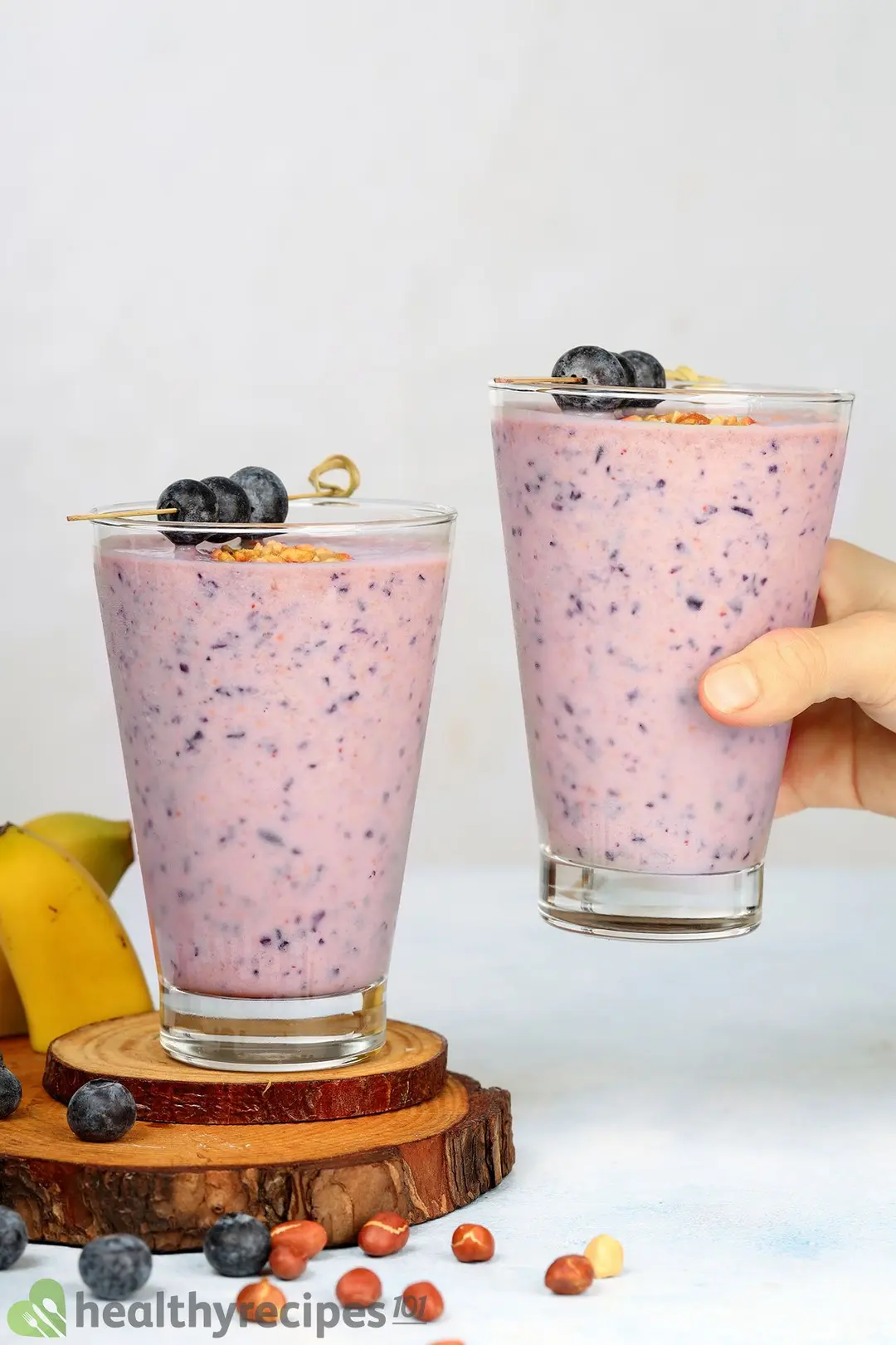 A glass of blueberry banana smoothie with crushed peanuts on two wooden coasters, with a blueberry smoothie about to be put next to it