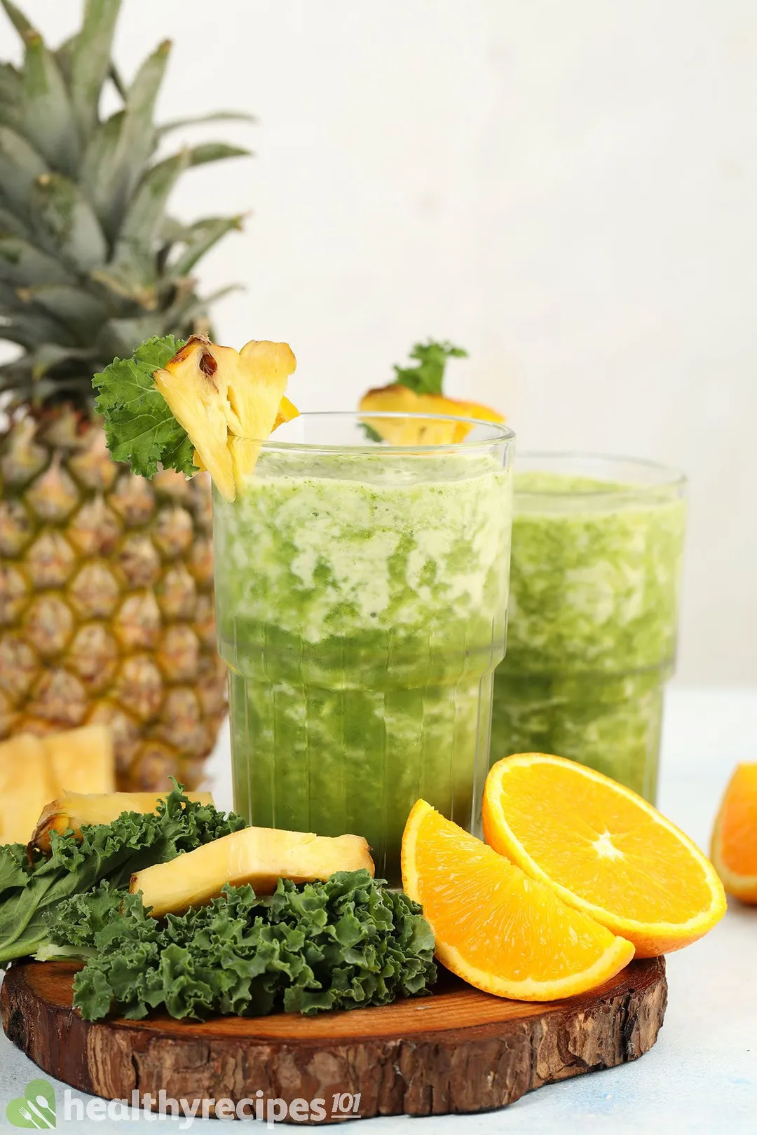 Two glasses of Pineapple Kale Smoothie placed on a wooden board near slices of orange, kale leaves, and a pineapple.