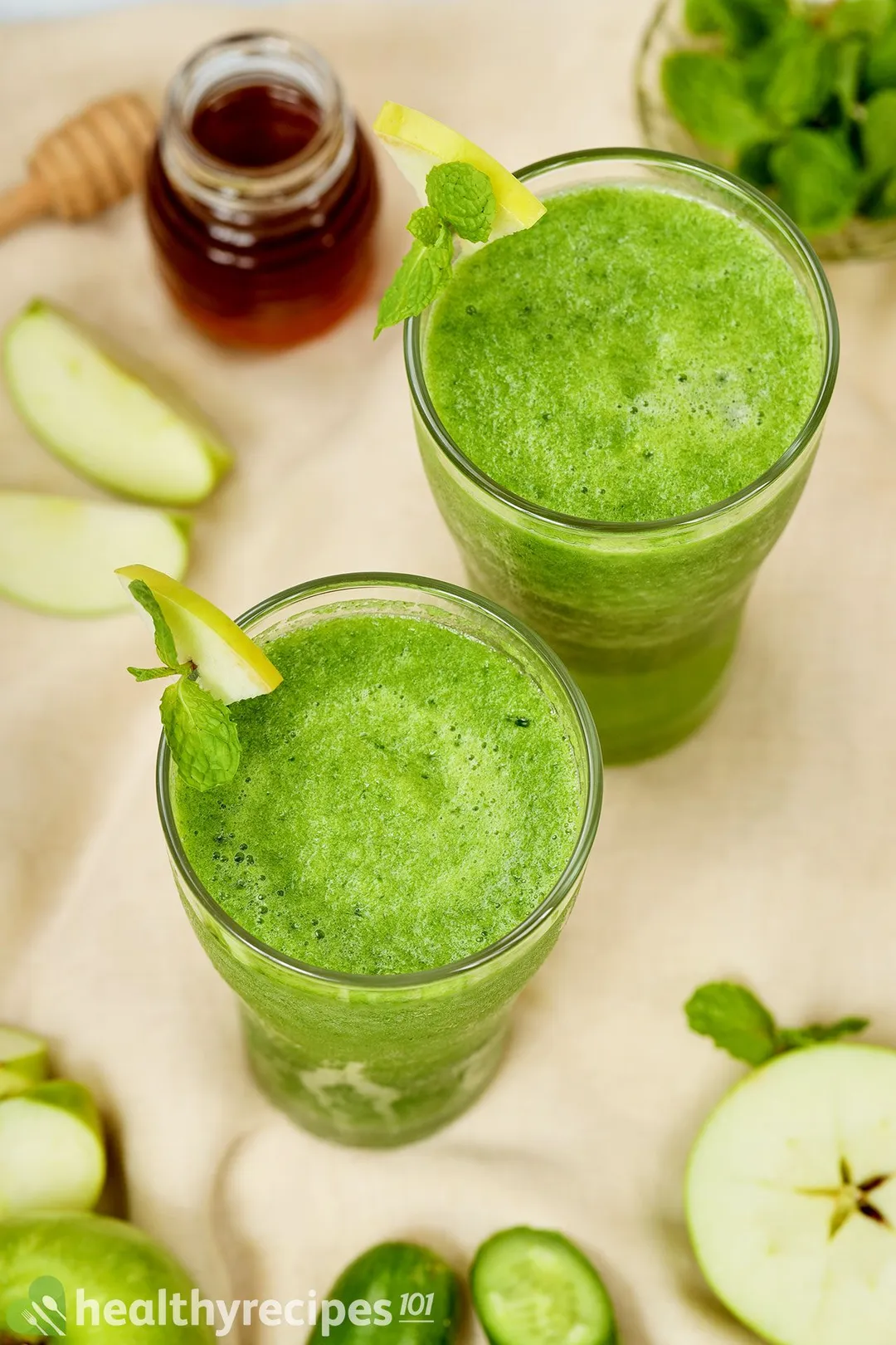 A high-angle shot of two sour apple smoothie glasses. Nearby is a small jar of honey and slices of green apples.