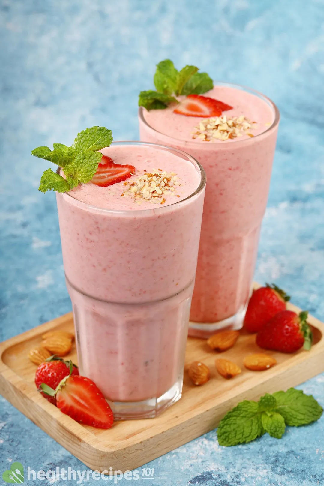 Two glasses of strawberry smoothie laid on a wooden board near strawberry slices and mint leaves.