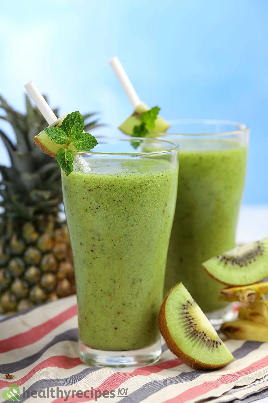 Two glasses of Pineapple Kiwi Smoothie placed on a tablecloth and in front of a pineapple.