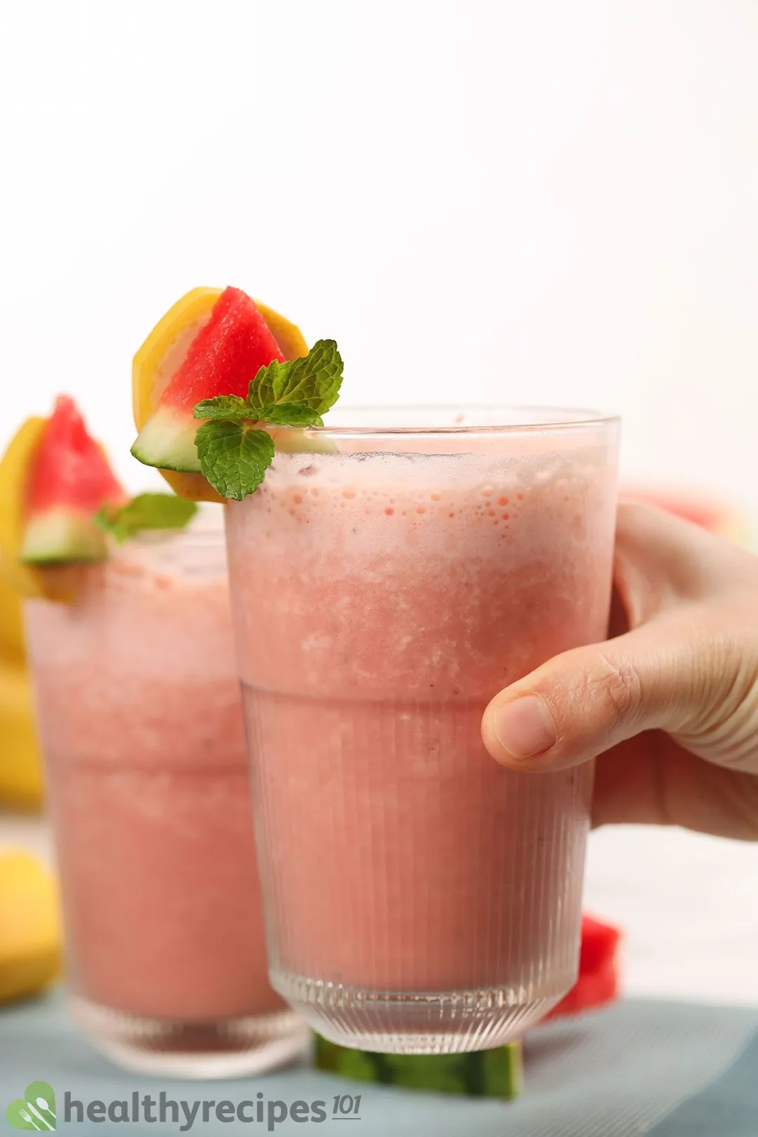 A hand picking up a glass of Watermelon Banana Smoothie placed in front of another identical glass.
