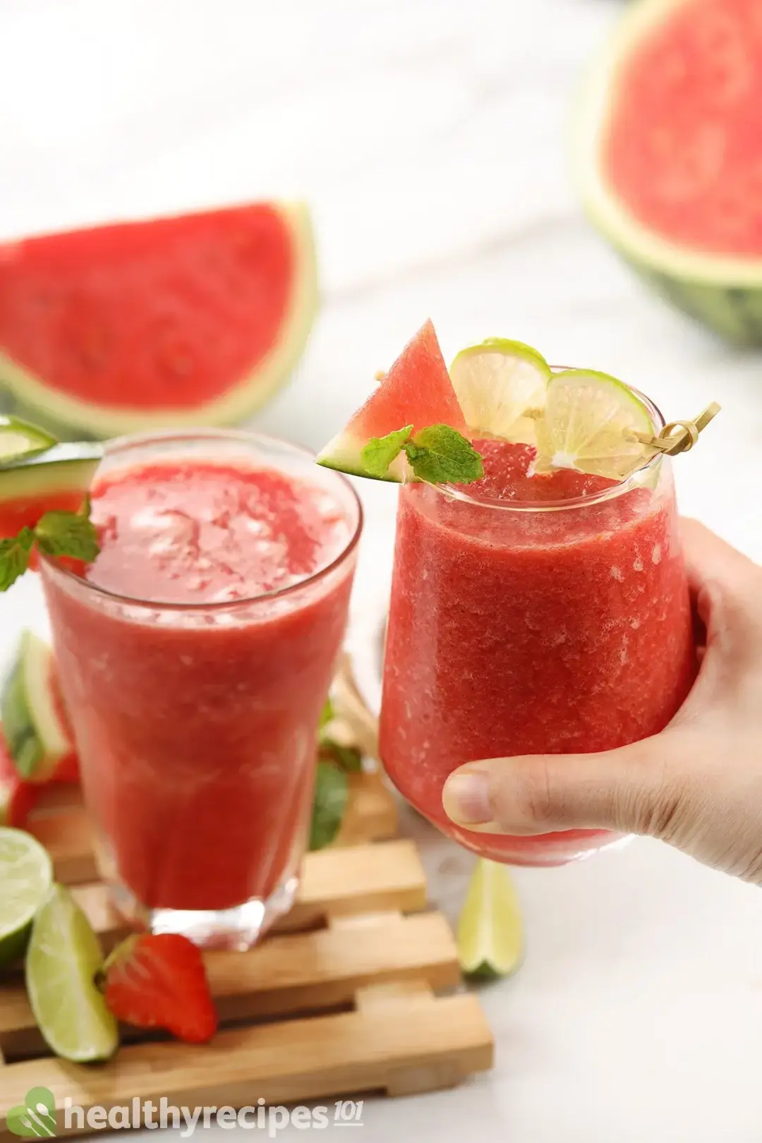 Two glasses of watermelon mojito smoothies, with one placed on a wooden tray surrounded with some fruit decorations and another held in the air by one hand