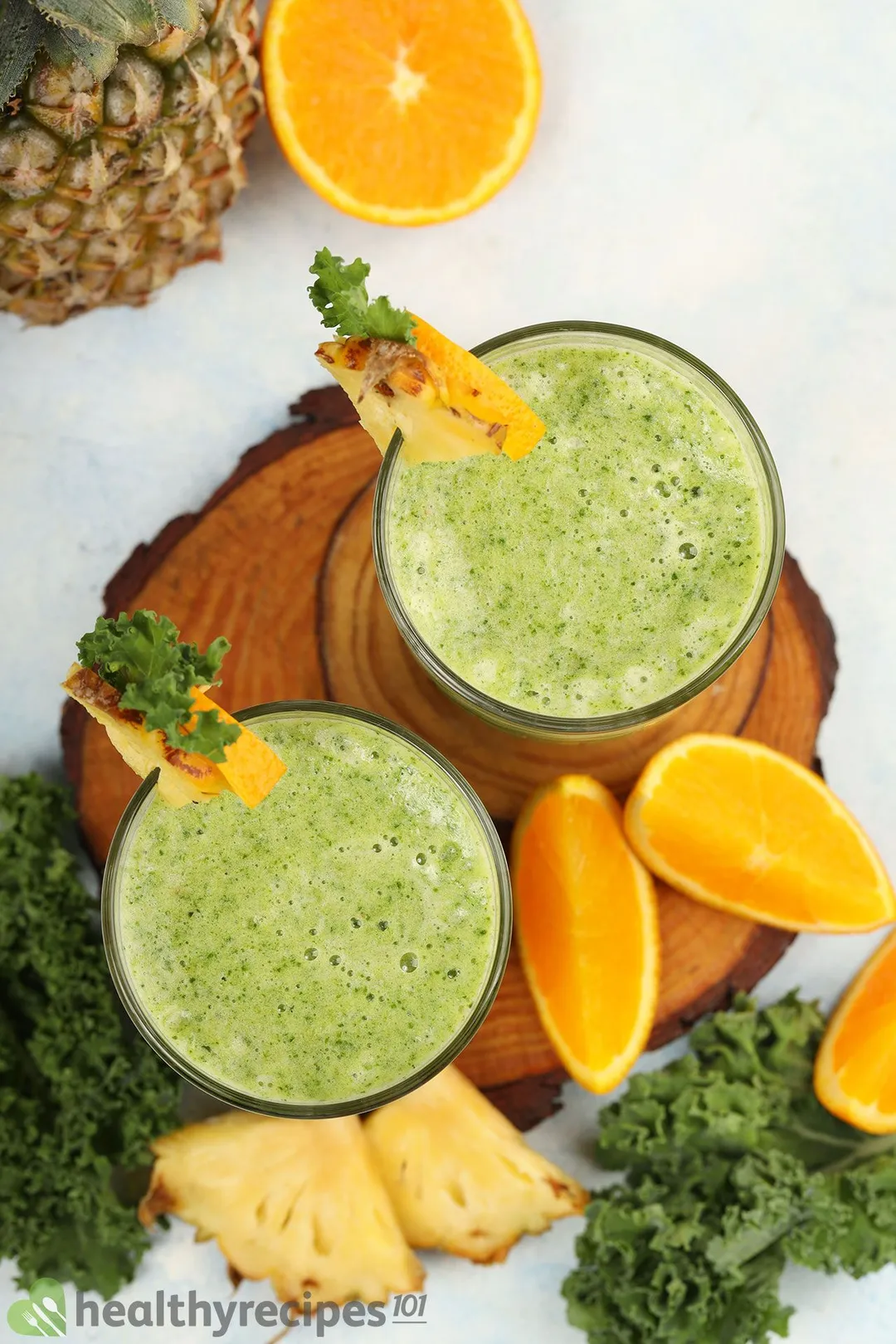 A high-angle shot of two Pineapple Kale Smoothie glasses, orange wedges, pineapple triangles, and kale leaves placed on a wooden board.