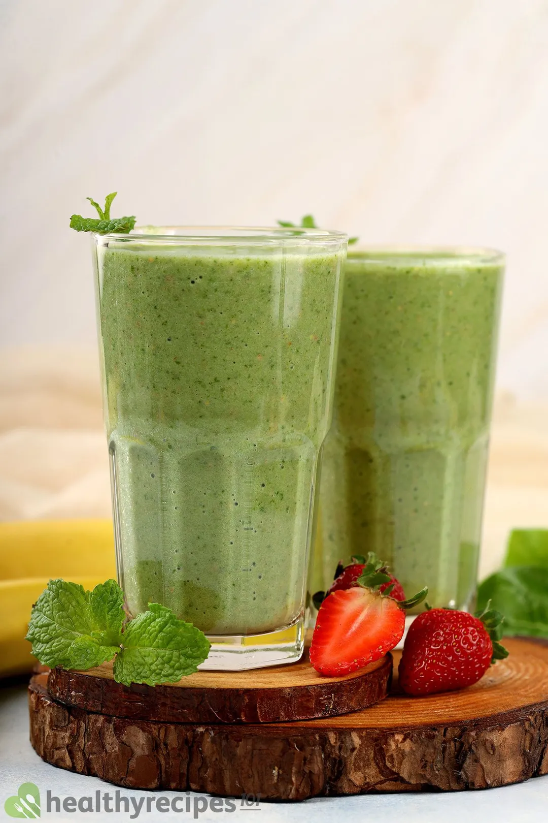 Two glasses of strawberry spinach banana smoothie placed on a wooden board and near mint leaves and strawberry slices.