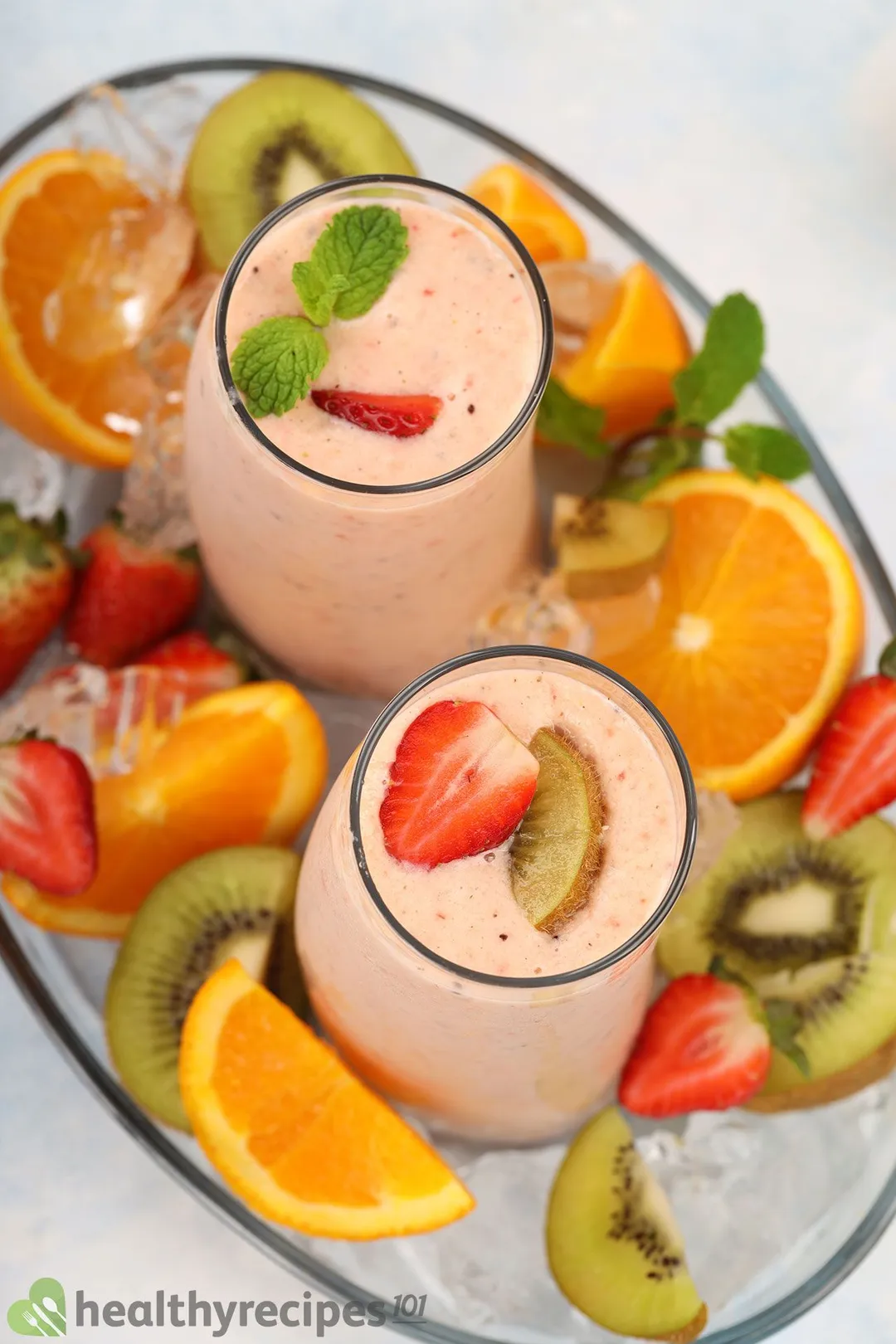 A high-angle shot of two glasses of kiwi quencher tropical smoothie placed in an ice bowl and surrounded by slices of kiwi, orange, and strawberry.