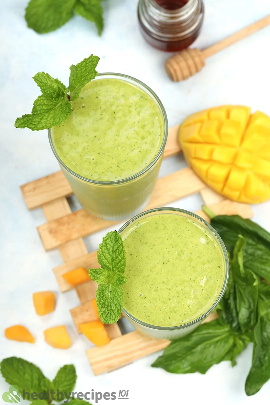 A high-angle shot of two glasses of Mango Spinach Smoothie placed on a wooden plank near a scored mango and fresh spinach leaves.
