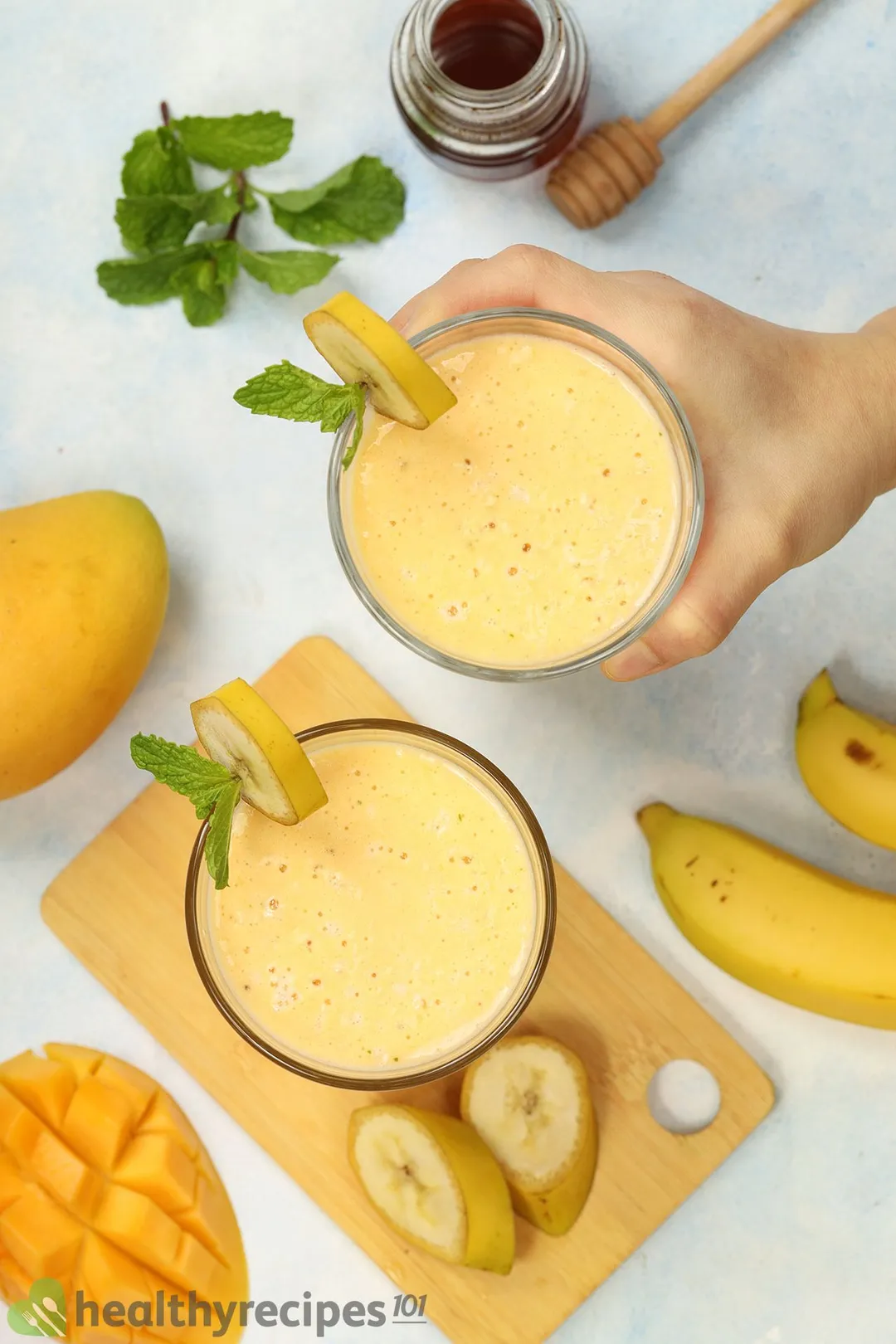 A high-angle shot of two glasses of Mango Banana Smoothie, bananas, a scored mango, mint leaves, and a small jar of honey.