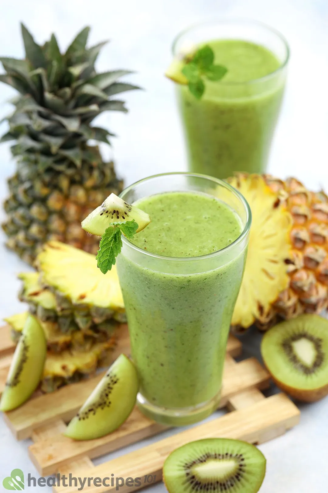 Two glasses of Pineapple Kiwi Smoothie placed on a wooden plank along with slices of pineapple and green kiwi.
