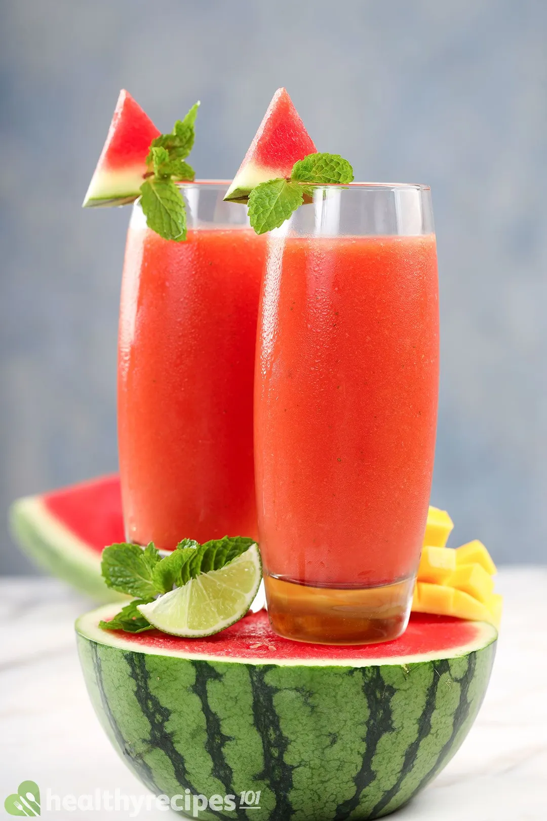 Two glasses of Mango Watermelon Smoothie, a lime wedge, and some mint leaves placed on half a watermelon.