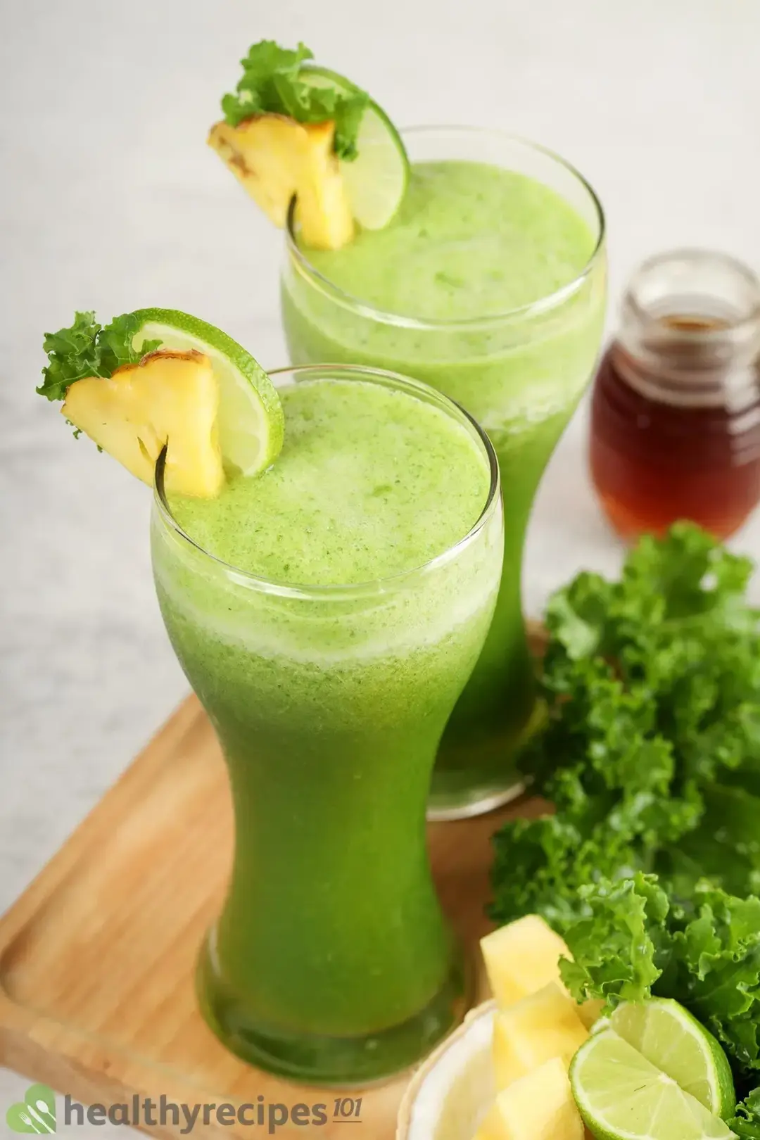 Two glasses of green smoothie next to each other decorated with kale, pineapple, and lime on a wooden board surrounded by other ingredients