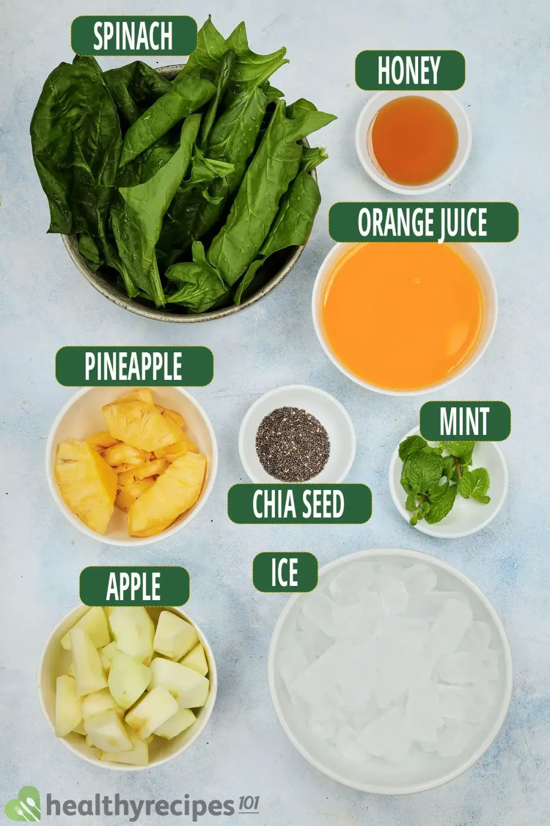 Ingredients needed for making a green apple smoothie, including bowls of apple cubes, pineapple slices, spinach leaves, spearmint leaves, chia seed, orange juice, and ice