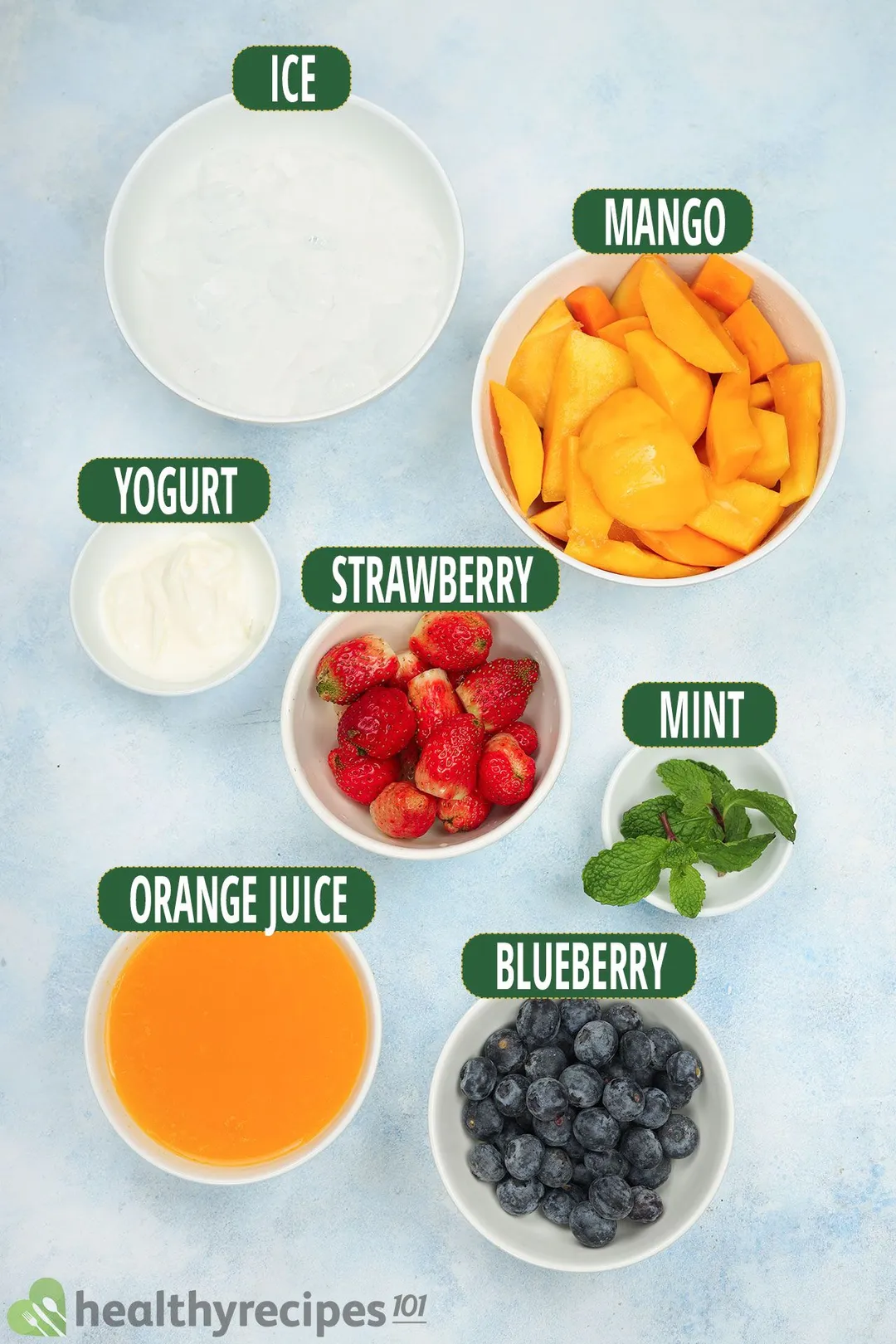Ingredients for Mango Berry Smoothie, including bowls of mango slices, blueberries, strawberries, mint leaves, orange juice, and more.