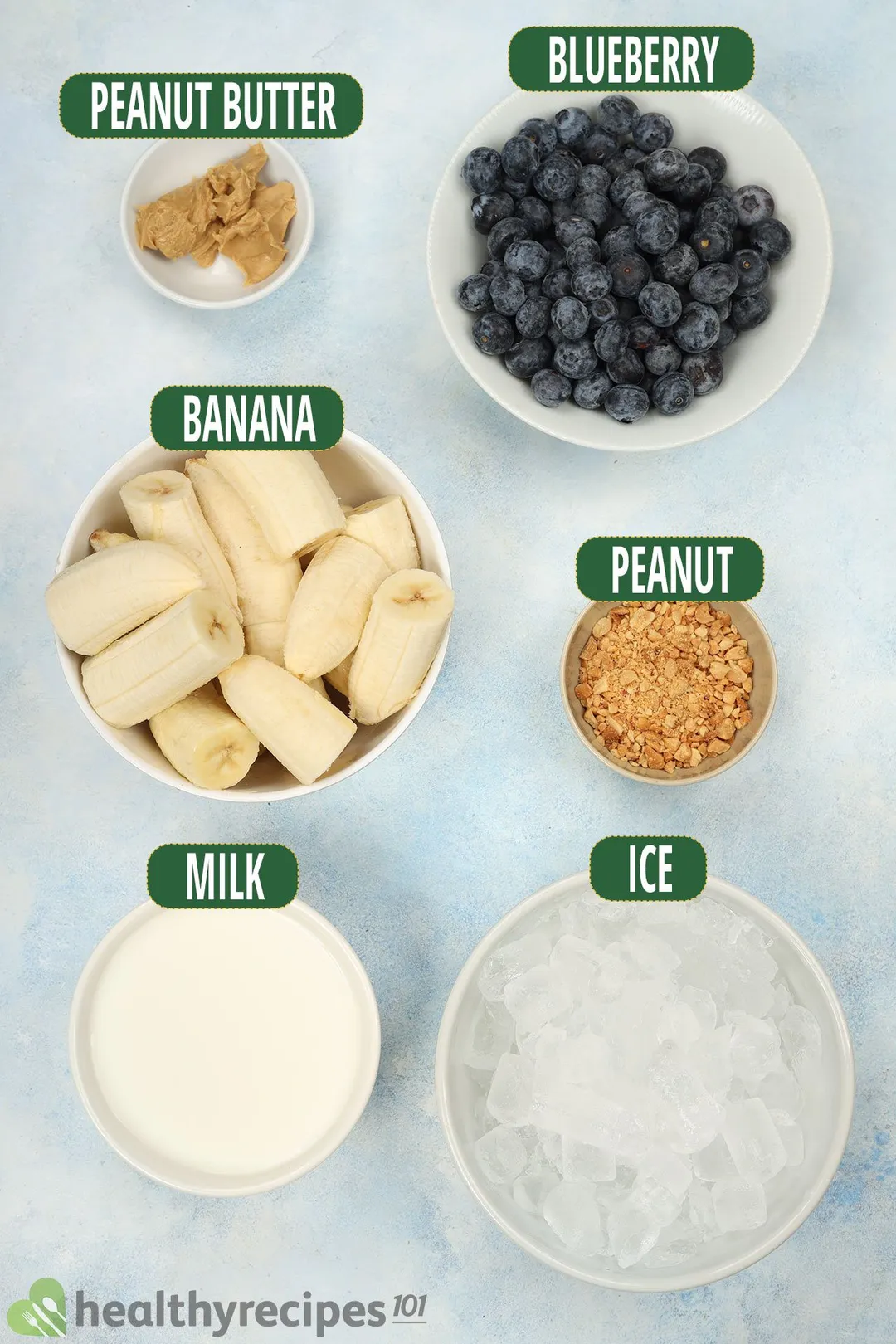 Ingredients in separate bowls: peanut butter, blueberries, cut bananas, crushed peanuts, milk, and ice nuggets