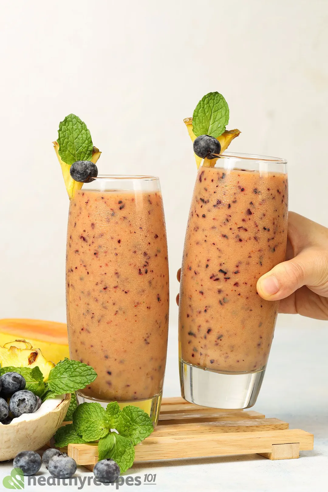 two glasses of mango blueberry smoothie, one on a tray and a hand holding the other
