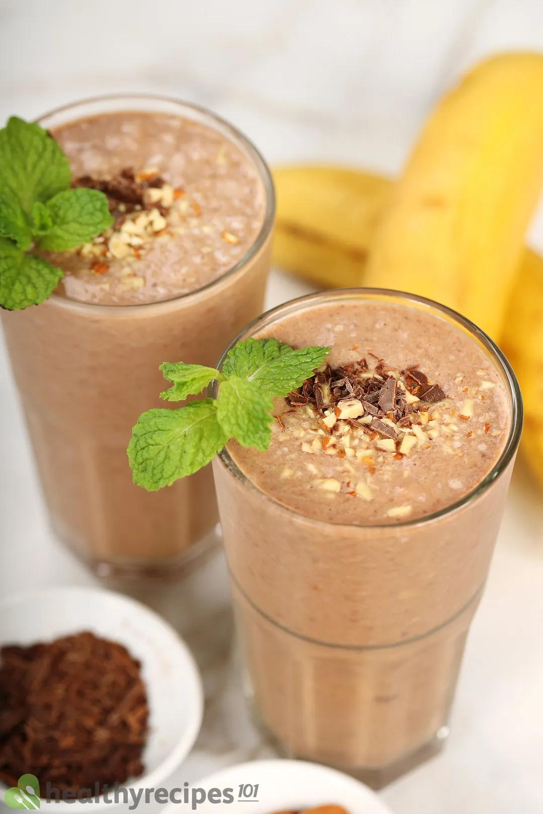 Two glasses of chocolate peanut butter banana smoothie placed near yellow unpeeled bananas and a small disk of minced chocolate.