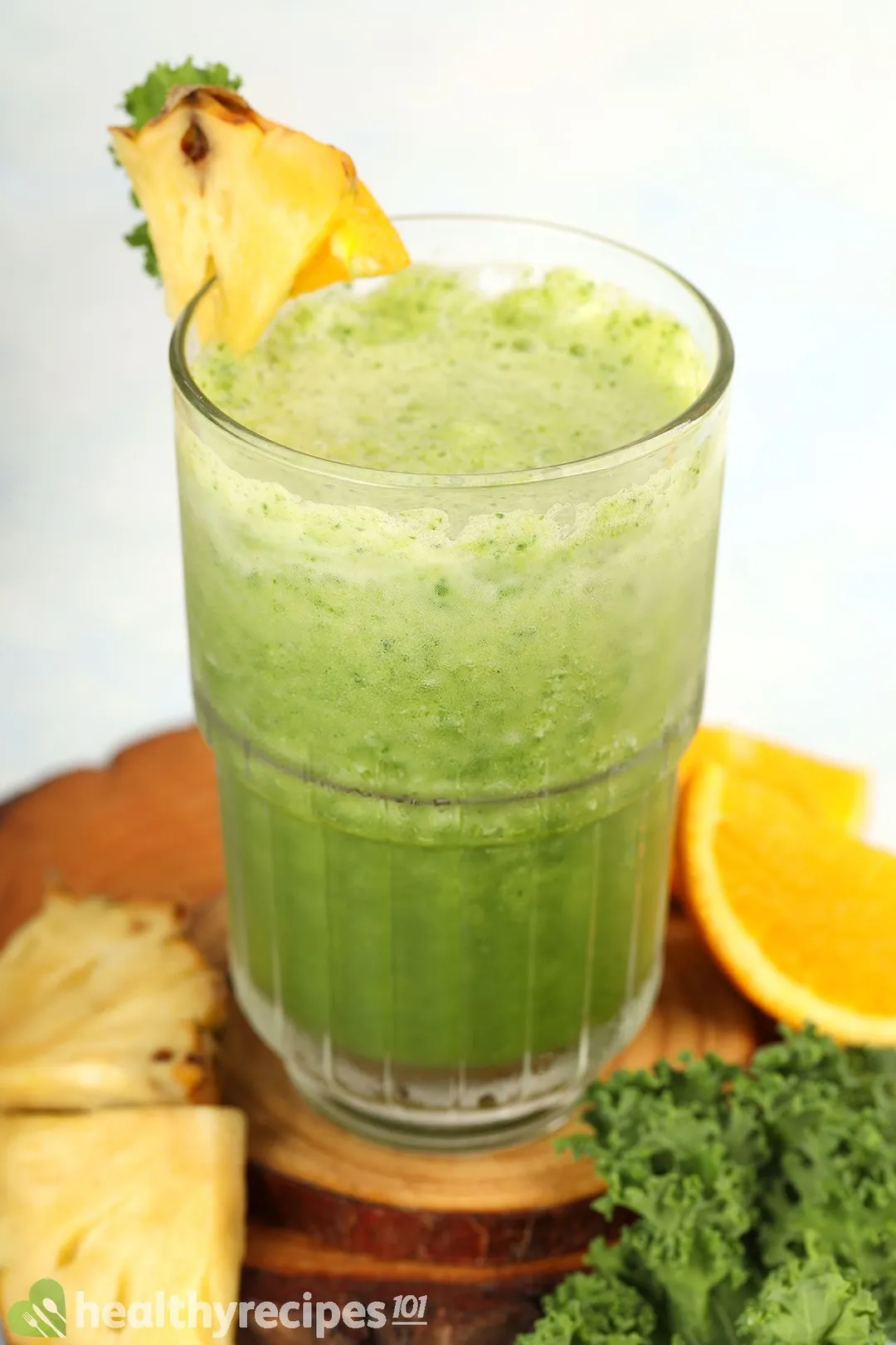 A glass of Pineapple Kale Smoothie placed on a wooden board near orange slices, pineapple triangles, and kale.