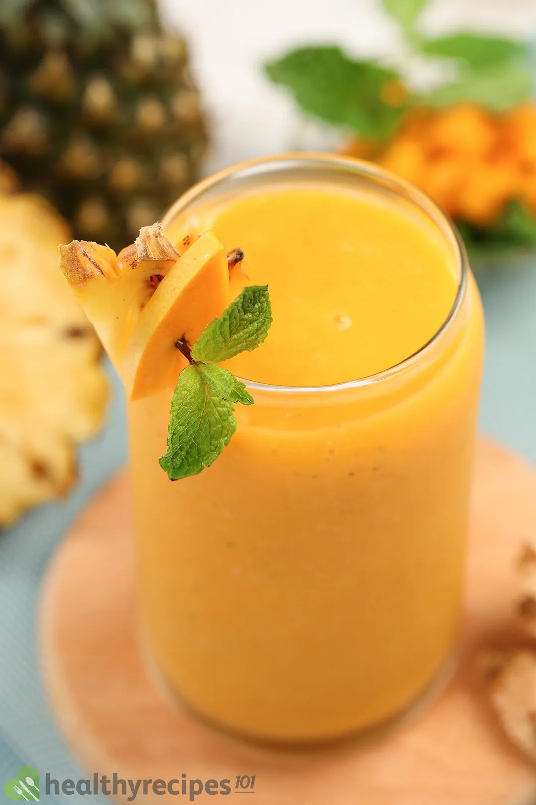 A glass of Mango Ginger Smoothie deocared with small slices of pineapples and a mint leaf placed on a wooden board.