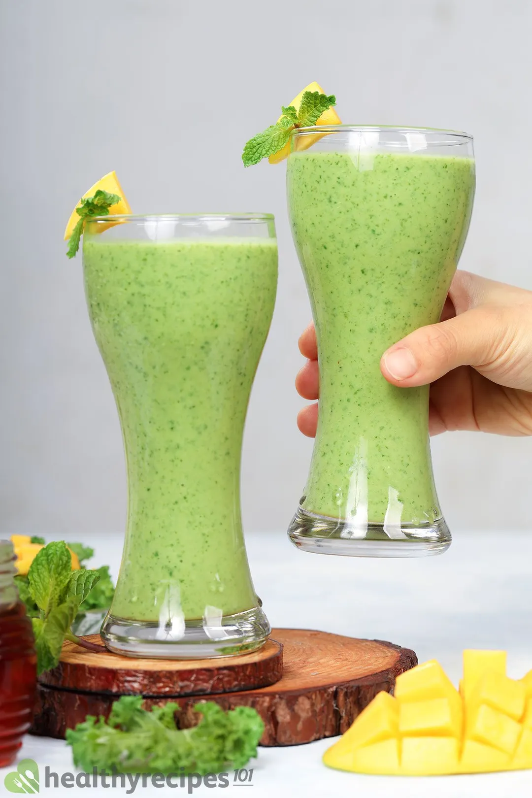 two glasses of mango kale smoothie, 1 on a tray and a hand holds the other