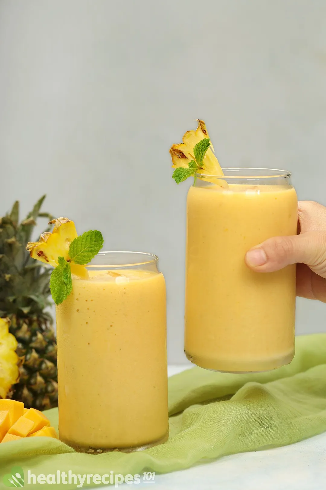 two glasses of mango pineapple, 1 on a table and hand holds the other